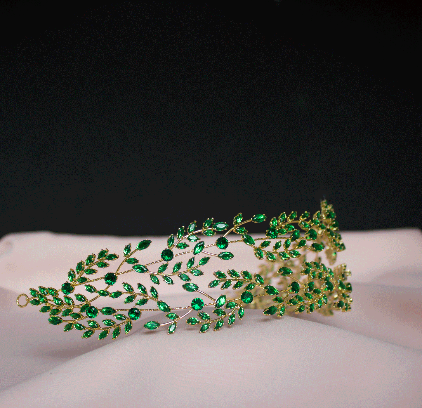 Elegante gouden haarband met een verfijnd ontwerp van takken en bladeren, versierd met sprankelende smaragdgroene kristallen, gepresenteerd op een zachte, lichtroze achtergrond.