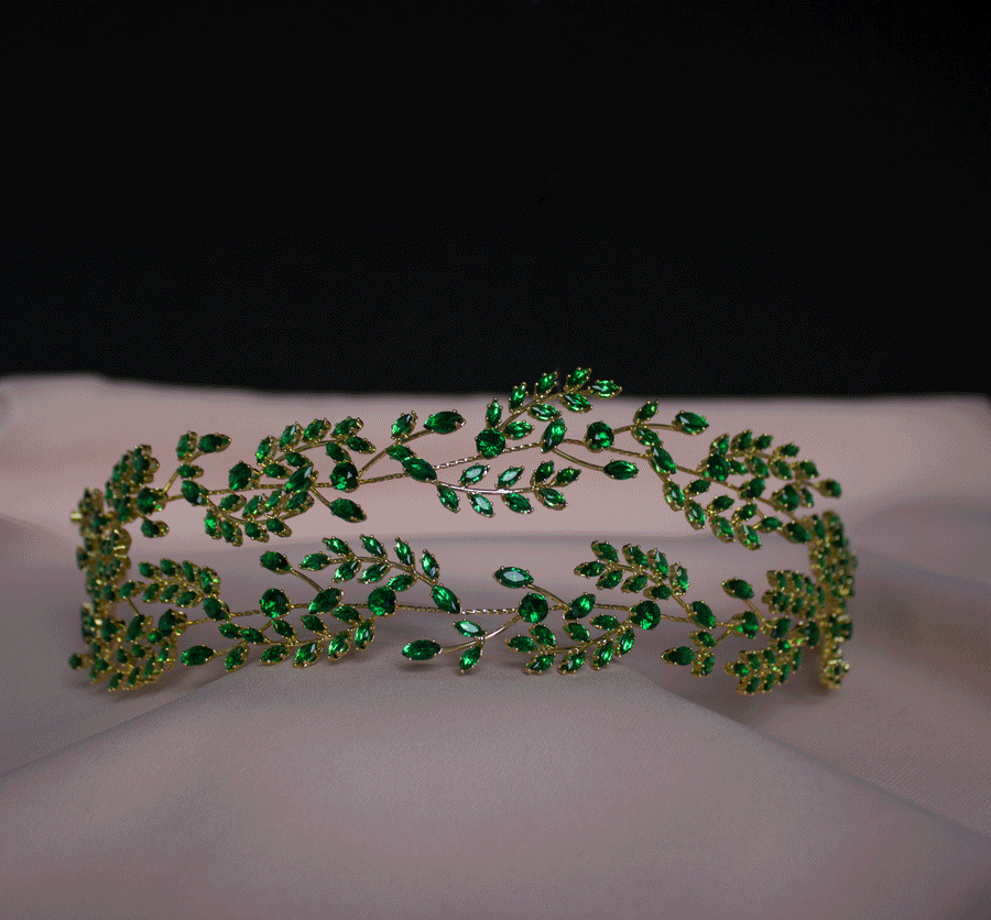 Elegante gouden haarband met een verfijnd ontwerp van takken en bladeren, versierd met sprankelende smaragdgroene kristallen, gepresenteerd op een zachte, lichtroze achtergrond.